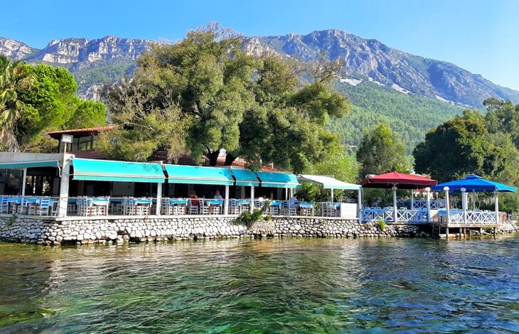 Akyaka Azmak Nehri kıyısında, dağ manzaralı yemyeşil ağaçların altında renkli tentelerle donatılmış nehir kenarı balıkçı restoranı.